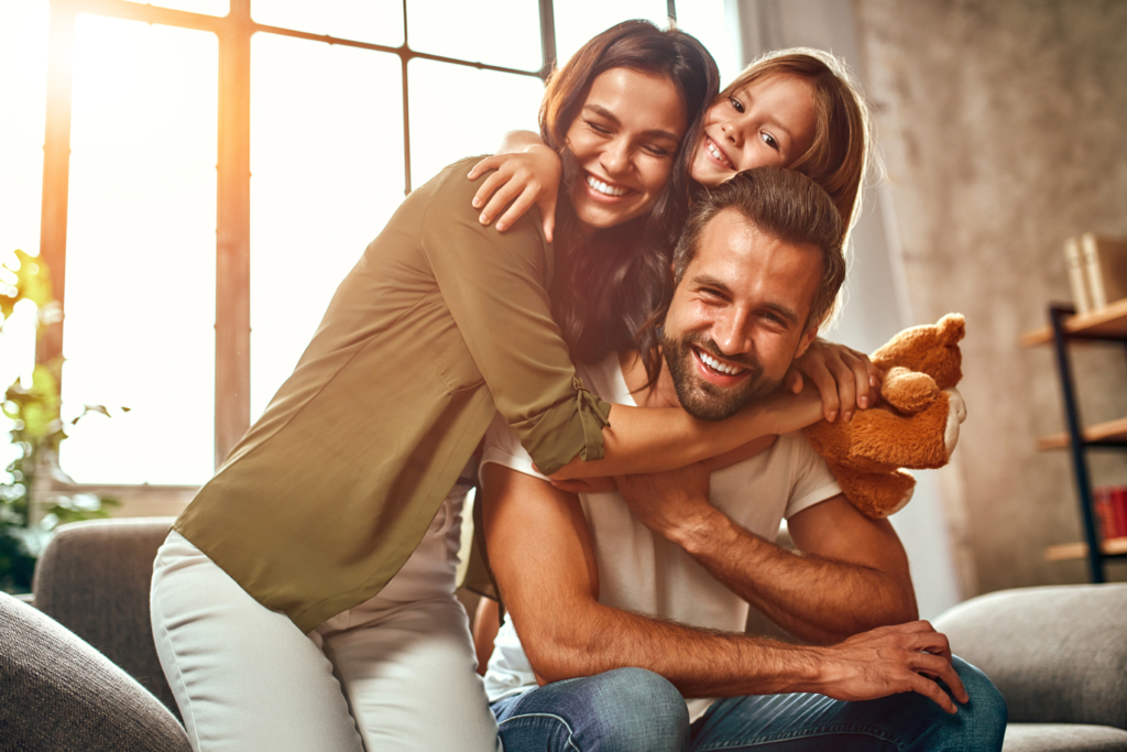 Glücklicher Vater und glückliche Mutter sitzen gemeinsam mit ihrer süßen Tochter und einem Teddybären auf dem Sofa im Wohnzimmer zu Hause, umarmen sich und haben Spaß.